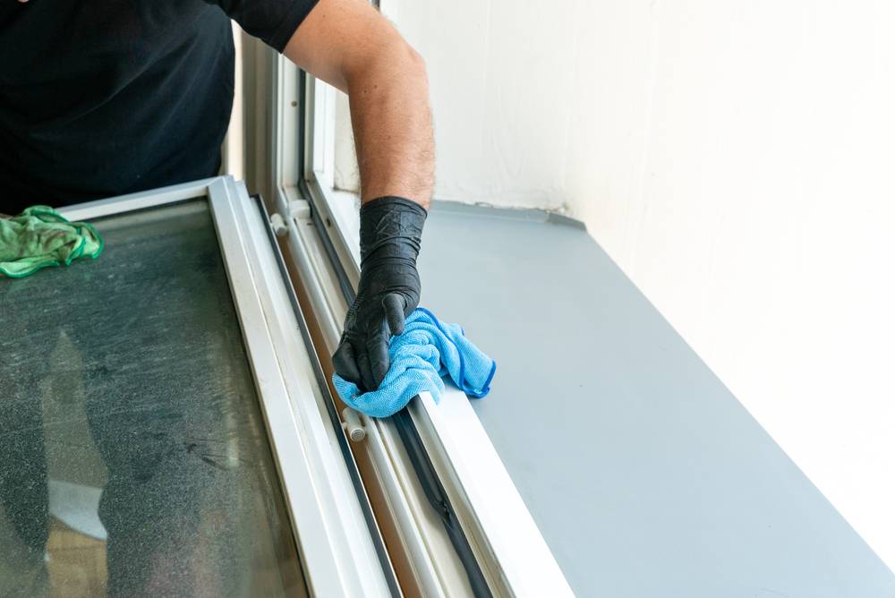 Hand im Schutzhandschuh reinigt einen Fensterrahmen mit einem Mikrofasertuch